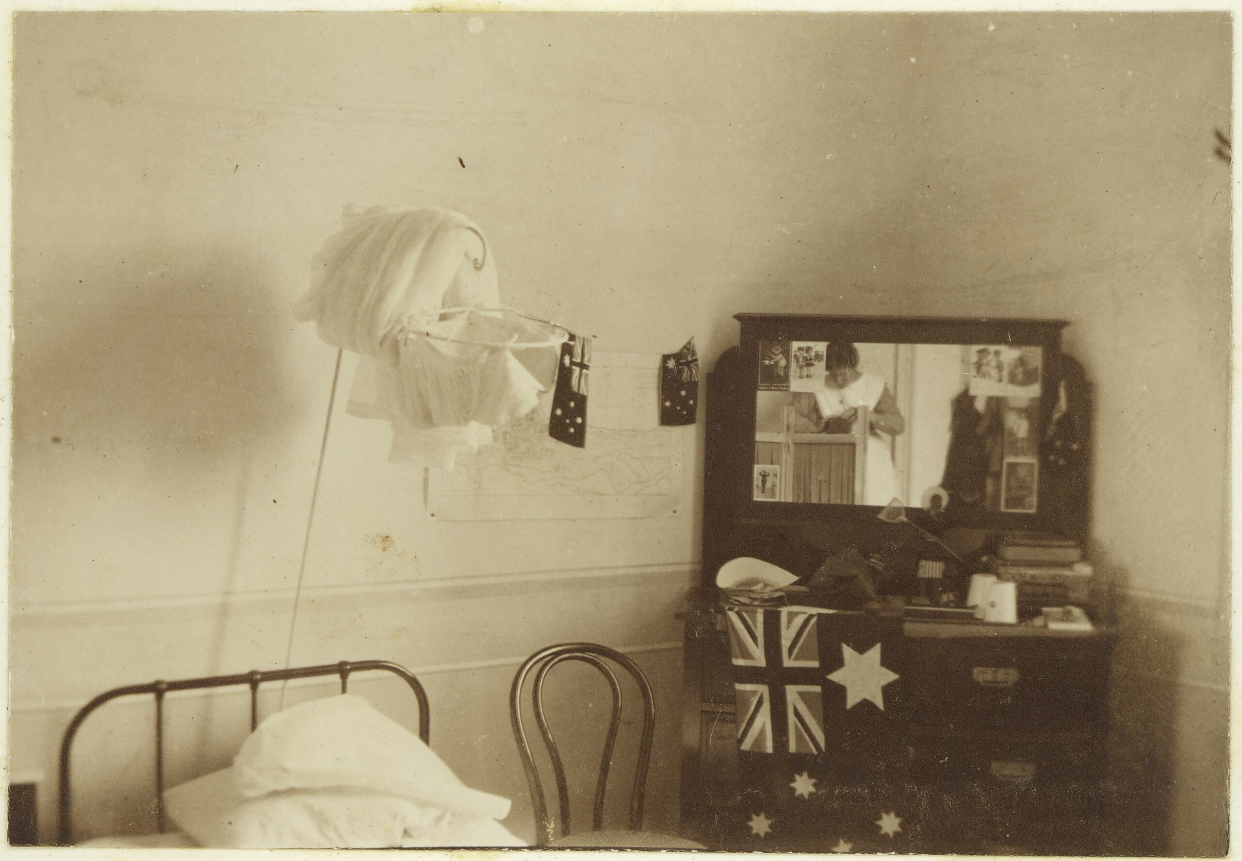 A small room is shown containing a single bed, dresser, mosquito net and several Australian flags hung on multiple surfaces. Assorted belongings are placed upon the dresser, and photos stuck to the mirror above it.