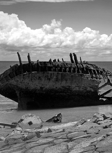 The broken hull of the Vyner Brooke is laying in a state of rusted decay in shallow water by the shoreline.