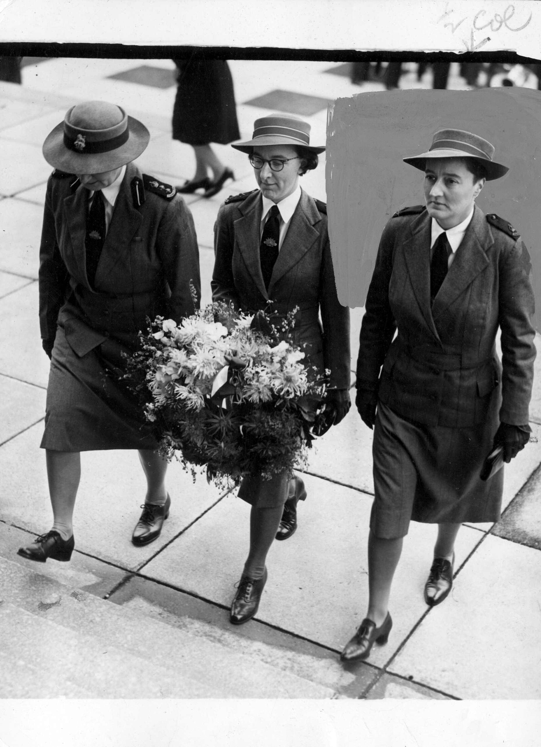 Three nurses in uniform walk in line with each other, approaching a set of stairs, with the middle nurse holding a wreath in her hands.