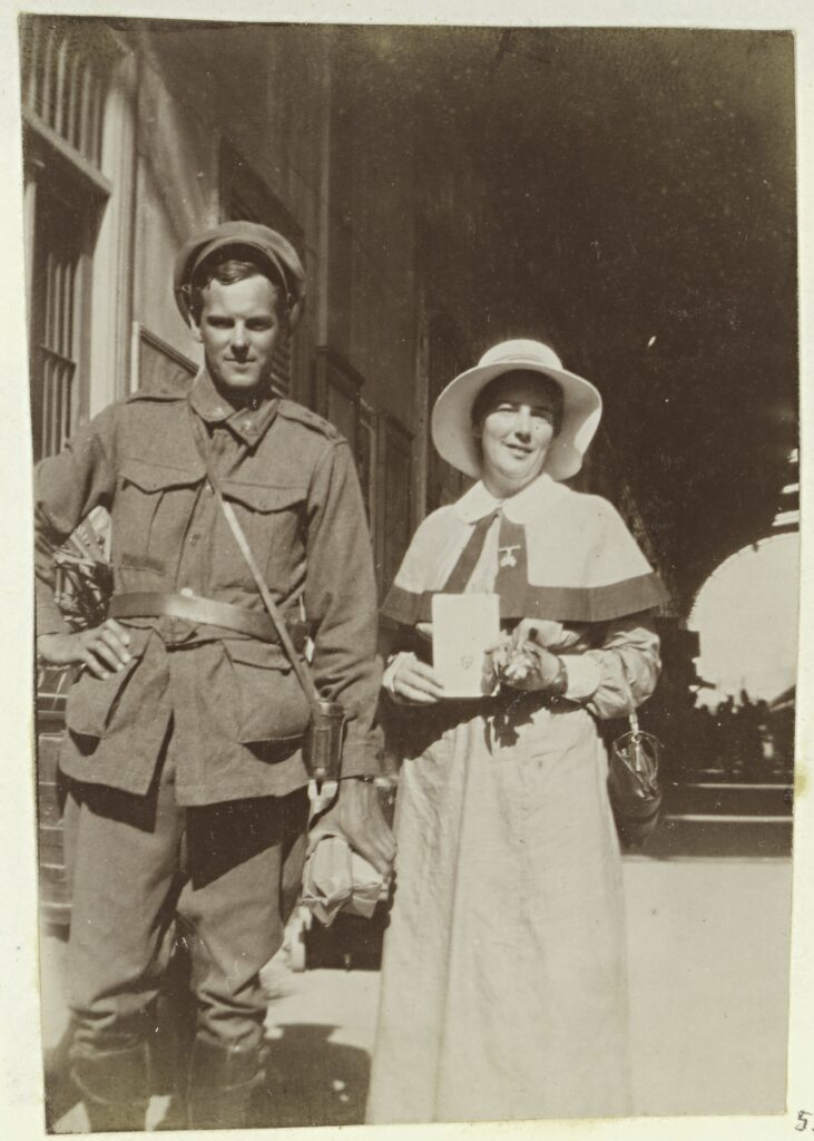 A soldier and nurse stand side by side, holding some purchased items in their hands, with a canopied street and urban buildings in the background behind them.