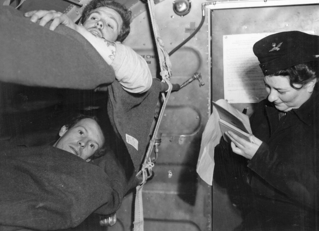 Two soldiers are lying down on canvas beds in an aircraft to the left of the image, with a nurse to their right writing down on a notepad and assessing them.